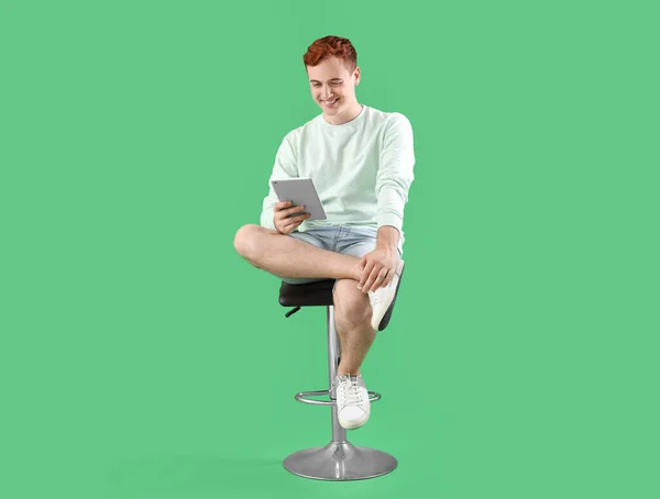 緑の背景に対して椅子に座っているタブレットを持つ幸せな若い赤毛の男 — ストック写真