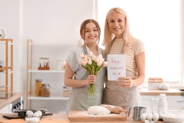 Genç bir kadın mutfakta annesini kart ve hediyeyle selamlıyor.