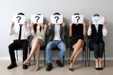 Genç başvuranlar, ellerinde kontrol kağıtları ve odada soru işaretleri olan kağıtlar tutuyorlar