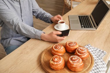 Elinde kahve ve cep telefonuyla mutfakta mesajlaşan genç adam, yakın plan.