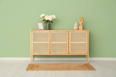 Yeşil duvarın yanındaki şifoniyerde fotoğraf çerçevesi ve biblo olan beyaz şakayıklar vazosu.