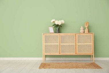 Yeşil duvarın yanındaki şifoniyerde fotoğraf çerçevesi ve biblo olan beyaz şakayıklar vazosu.