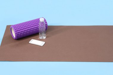 Köpük silindirli spor paspası, cep telefonu ve mavi arka planda bir şişe su.