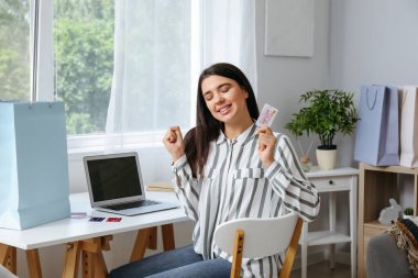 Evde hediye kartı ve laptop alışverişi yapan mutlu genç bir kadın.