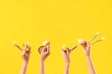 Sarı arka planda makyaj fırçası, sünger ve pudra olan kadınlar