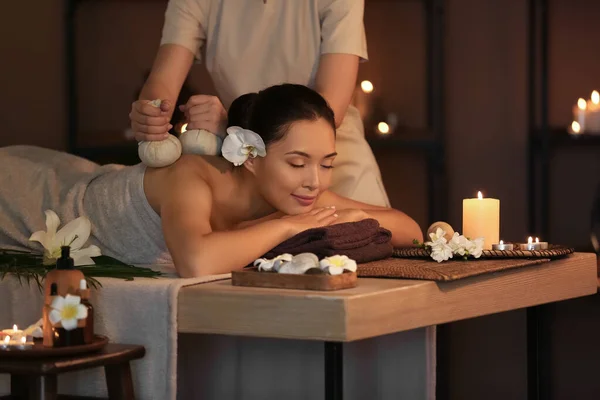 Young woman undergoing treatment with herbal bags in dark spa salon