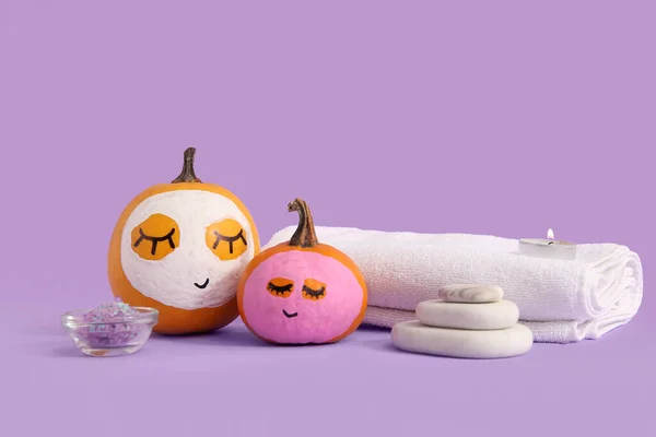 Pumpkins with masks and spa supplies on lilac background