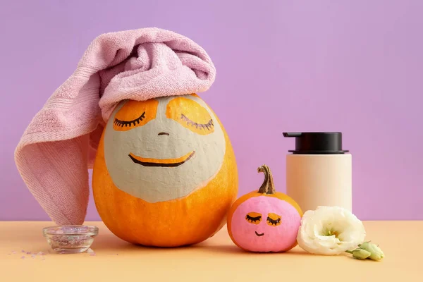 Pumpkins with masks, spa supplies and flowers on table against lilac background