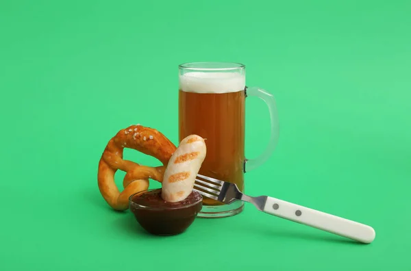 stock image Fork with tasty Bavarian sausage, pretzel, sauce and mug of beer on green background. Oktoberfest celebration