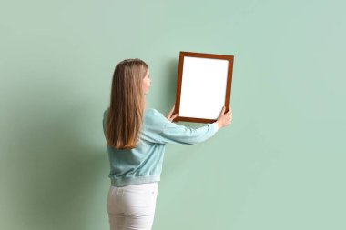 Evdeki yeşil duvara boş bir çerçeve asan genç kadın.