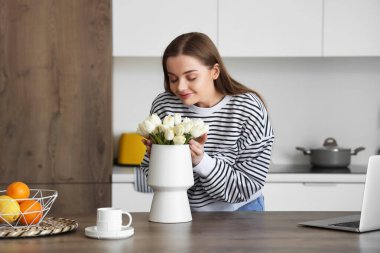 Mutfakta beyaz lale çiçekleriyle vazo tutan güzel genç bir kadın.