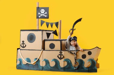 Korsan gibi giyinmiş şirin küçük kız sarı arka planda karton gemide kılıçla oynuyor.