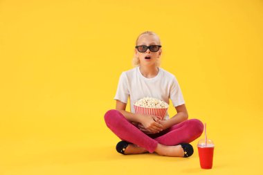 Üç boyutlu sinema gözlüklü, elinde patlamış mısır ve kolayla sarı arka planda oturan şok olmuş bir kız.