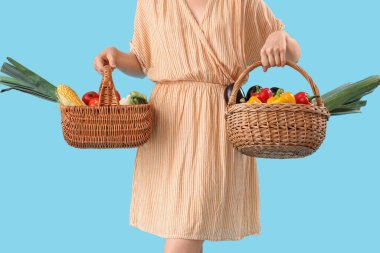 Mavi arka planda farklı olgun sebzelerle dolu sepetleri olan genç kadın çiftçi, yakın plan.