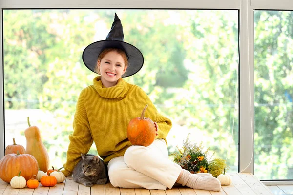 Young woman in witch hat with cute cat and Halloween pumpkins near window at home