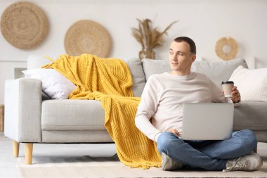 Evde bir fincan kahve ve dizüstü bilgisayarı olan genç bir adam var.