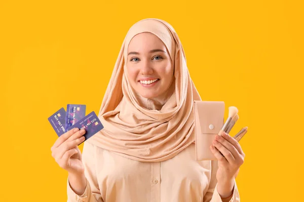 Young Muslim woman with credit cards and makeup brushes on yellow background