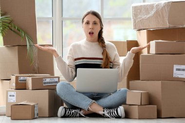 Evde paketler ve laptopla şok olmuş genç bir kadın var.