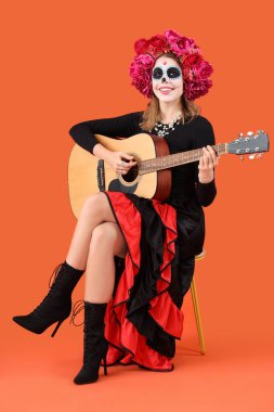 Kafatası boyalı genç bir kadın turuncu arka planda gitar çalıyor. Meksika Ölüler Günü (El Dia de Muertos) kutlamaları