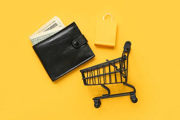 Small shopping cart, bag and wallet with money on yellow background