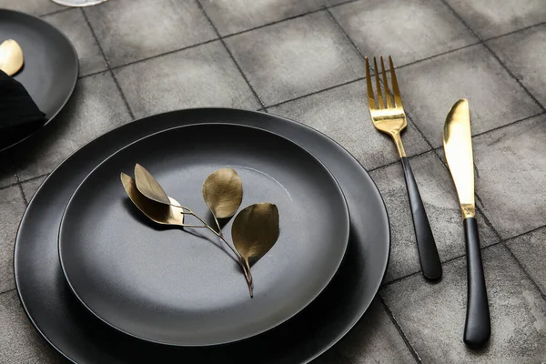 Elegant table setting with black plates, golden leaves and cutlery on grey tiled background