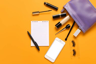 Makyaj malzemeli makyaj çantası, modern cep telefonu ve turuncu arka planda not defteri.