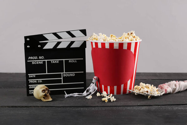 Bucket with popcorn, Halloween decor and movie clapperboard on wooden table against grey background