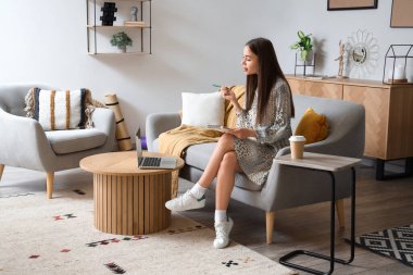 Dizüstü bilgisayarı olan genç bir kadın evdeki kanepeye notlar yazıyor.