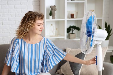 Oturma odasındaki kanepede elektrikli yelpazeli güzel, mutlu genç bir kadın oturuyor.