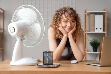İş yerinde elektrikli yelpazesi olan güzel, mutlu genç bir kadın.