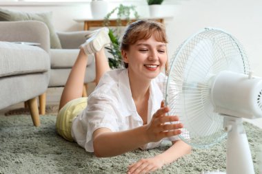 Evde yerde yatan, elinde vantilatör olan genç bir kadın.