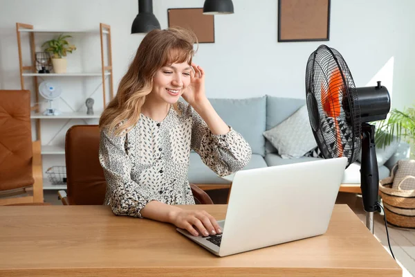 Beautiful happy young woman with laptop and electric fan sitting at table in living room