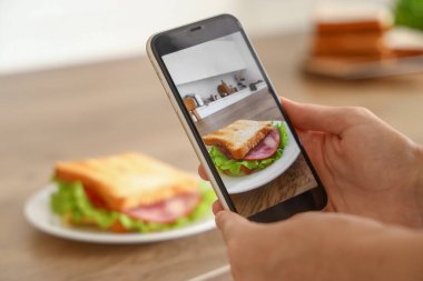 Cep telefonlu genç bir kadın mutfakta lezzetli bir sandviçin fotoğrafını çekiyor.