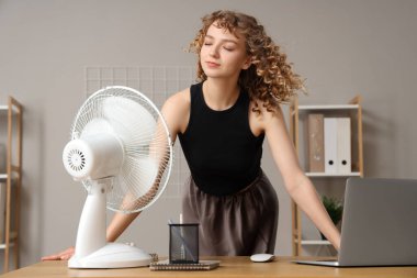 İş yerinde elektrikli yelpazesi olan güzel, mutlu genç bir kadın.