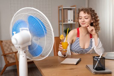 Elektrikli yelpazesi ve kokteyli olan güzel, mutlu genç bir kadın iş yerinde sandalyede oturuyor.