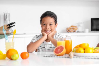 Küçük Afro-Amerikalı çocuk taze meyve suyu ve farklı tabaklar ile mutfaktaki masada oturuyor.