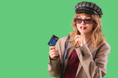 Yeşil arka planda sessizlik jesti gösteren kredi kartı olan şık genç bir kadın.