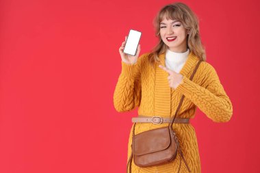 Kırmızı arka planda cep telefonuna işaret eden şık genç bir kadın.