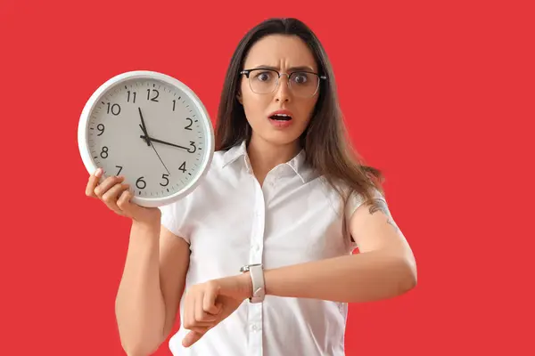 Stressed young woman with clock and wristwatch on red background. Deadline concept