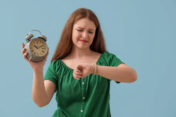 Stressed young woman with alarm clock looking at wristwatch on blue background. Deadline concept