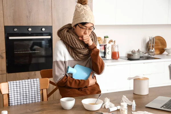 Ill Asian man with hot water bottle in kitchen