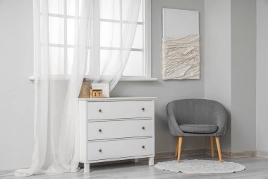 Açık renkli oturma odası, pencerenin yanındaki gri koltuk ve çekmeceler.