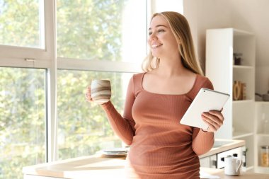 Mutfakta bir fincan sıcak çay ve tablet bilgisayarlı genç bir kadın.