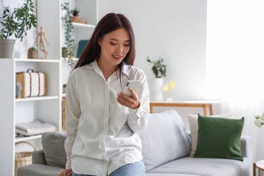 Oturma odasında cep telefonu kullanan güzel bir Asyalı kadın.