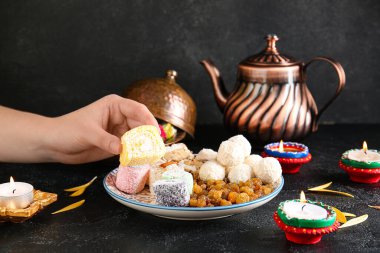 Elinde diya lambası, mumlar, çaydanlık ve siyah arka planda farklı ikramlar olan kadın eli. Divaly kutlaması