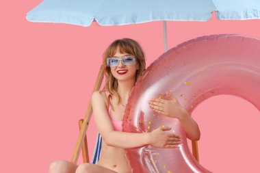 Plaj şemsiyesi ve pembe arka planda yüzme yüzüğü olan güverte sandalyesinde oturan genç bir kadın.