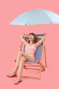 Güverte sandalyesinde pembe arka planda plaj şemsiyesi olan genç bir kadın.