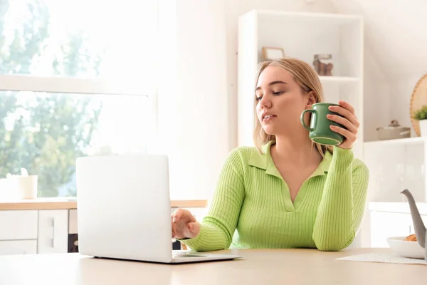 Young woman with cup of hot tea using laptop at table in kitchen