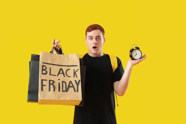 Shocked young redhead man with shopping bags and alarm clock on yellow background. Black Friday sale