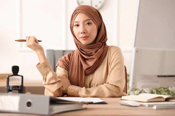 Beautiful young Asian woman in hijab with pen sitting at office